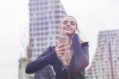 Akıllı telefonları ve kulaklıkları olan, açık havada müzik dinleyen gülümseyen genç bir kadın.