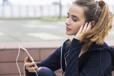 Akıllı telefonları ve kulaklıkları olan, açık havada müzik dinleyen gülümseyen genç bir kadın.