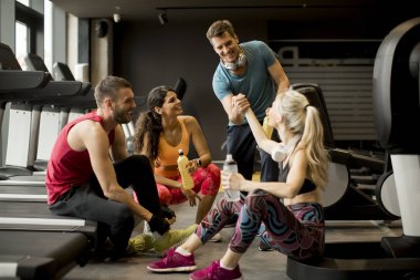 Spor giyimli bir grup genç, antrenman dan sonra spor salonunun zemininde otururken birlikte konuşup gülüyor