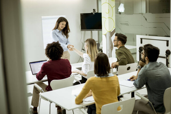 Smiling young woman standing near whiteboard and shaking hand to her female colleague in the small startup office