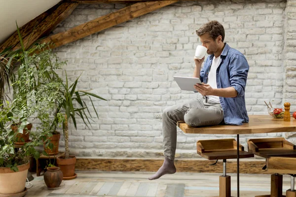 Yakışıklı genç adam dijital tablet kullanarak ve rustik odada masada otururken kahve içme