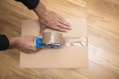 Bir nakliye kutusunu mühürlemek için bant dağıtıcısı kullanan bir adamın elleri