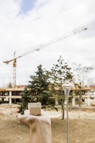 Hand holding small house model in front of construction site, re — Stock Photo, Image