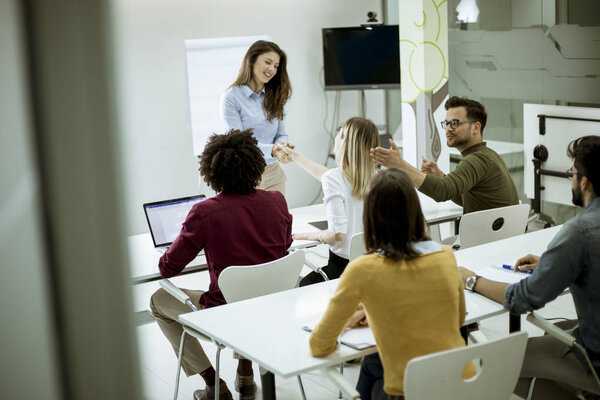 Smiling young woman standing near whiteboard and shaking hand to her female colleague in the small startup office