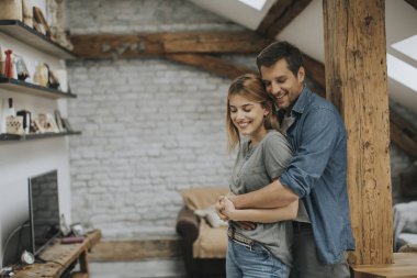 Genç bir adam ve bir kadın evlerinde durup birbirlerine sarılırlar ve kocaları karısına nazikçe sarılırlar.