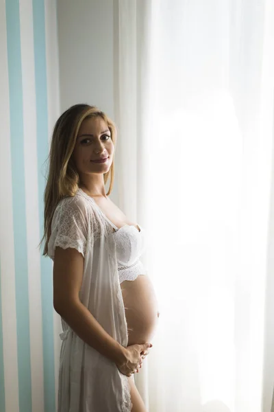 漂亮的年轻孕妇站在窗口在蓝色剥离墙在房间里在白天 — 图库照片
