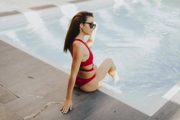 身着红色比基尼 戴着太阳镜的迷人年轻女子坐在池畔 享受夏日的阳光 — 图库照片