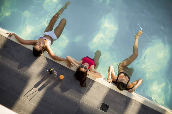 三个美丽的苗条年轻女子在比基尼放松和喝鸡尾酒在游泳池的池畔 — 图库照片