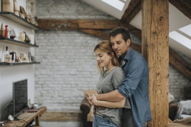 Genç bir adam ve bir kadın evlerinde durup birbirlerine sarılırlar ve kocaları karısına nazikçe sarılırlar.