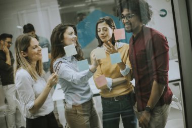 Genç iş adamları grubu başlangıç ofisinde post it notlar ve çıkartmalar kullanarak cam duvar önünde tartışıyor