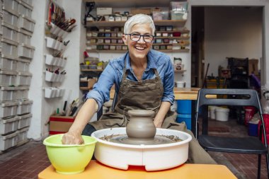 Onun atölyesinde otururken çömlek tekerleği üzerinde çalışan iyi görünümlü yaşlı kadın çömlekçi