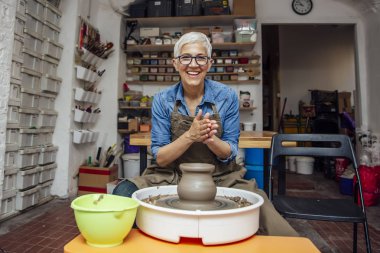 Onun atölyesinde otururken çömlek tekerleği üzerinde çalışan iyi görünümlü yaşlı kadın çömlekçi