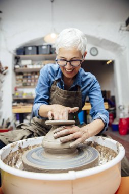 Onun atölyesinde otururken çömlek tekerleği üzerinde çalışan iyi görünümlü yaşlı kadın çömlekçi