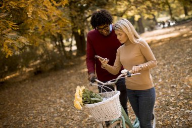 Güz parkında bisiklet süren ve cep telefonu kullanan çok ırklı genç bir çift.