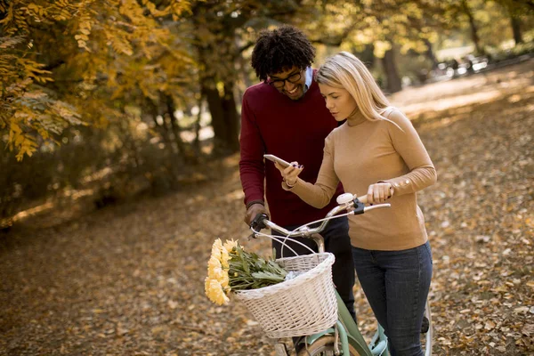 Güz parkında bisiklet süren ve cep telefonu kullanan çok ırklı genç bir çift.