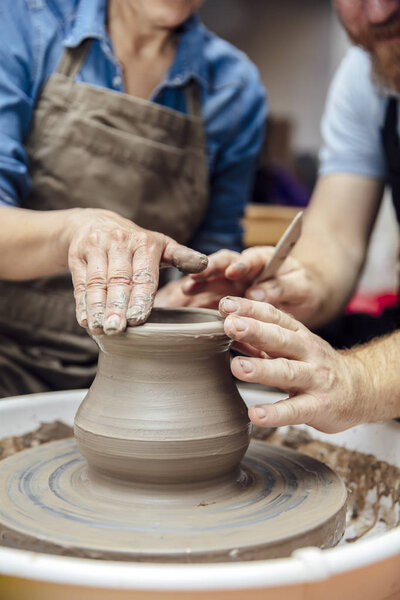 Senior woman spinning clay on a wheel with a help of a teacher at pottery class