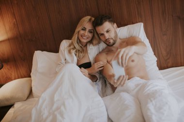 Mutlu çift evde yatakta uzanıyor ve cep telefonu kullanıyor.