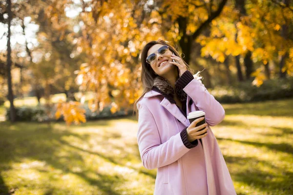 明るい公園で彼女の携帯電話を使用してピンクのコートを着て陽気な若い女性と飲み物を取るコーヒー — ストック写真