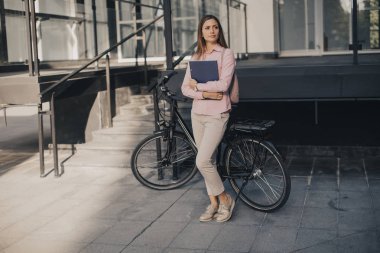 Young woman with files in the  hands standing outdoor in front of electric bike