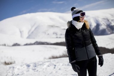 Genç bir kadın gündüz vakti kayak elbisesiyle karlı bir dağın yamacında duruyor.