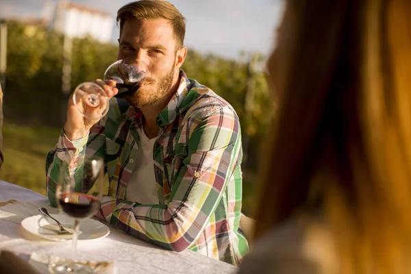 一个英俊的年轻人坐在桌旁 日落时在葡萄园里喝着红酒 — 图库照片