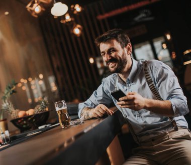 Mutlu genç adam bira içiyor, sigara içiyor ve barda cep telefonu kullanıyor.