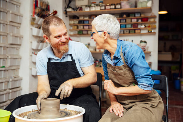 Senior woman spinning clay on a wheel with a help of a teacher at pottery class