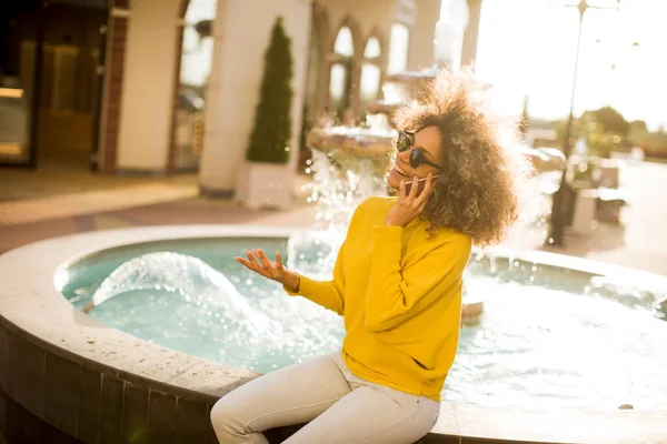 Attractive young woman with curly hair using her touch screen mobile cell phone by the fountain