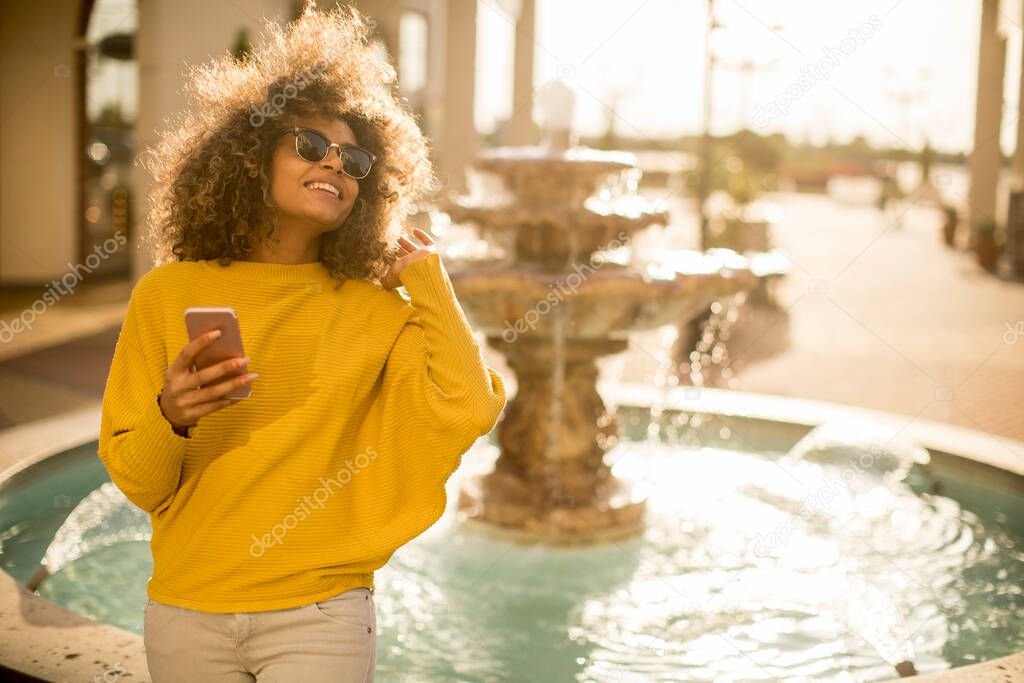 Attractive young woman with curly hair using her touch screen mobile cell phone by the fountain