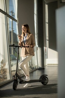 Ofis penceresinin yanında elektrikli scooterlı ve cep telefonu kullanan güzel genç bir kadın.
