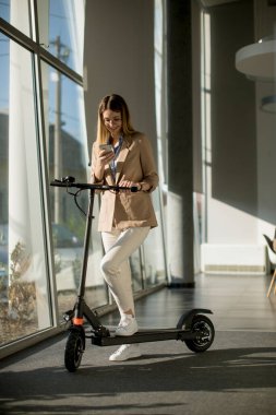 Ofis penceresinin yanında elektrikli scooterlı ve cep telefonu kullanan güzel genç bir kadın.