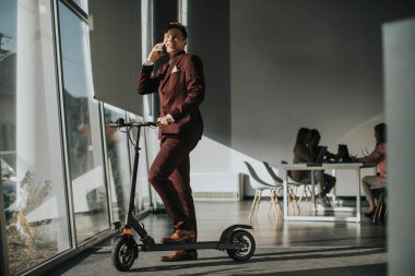 Ofis penceresinin yanında elektrikli scooterlı yakışıklı ve cep telefonu kullanan genç bir işadamı.