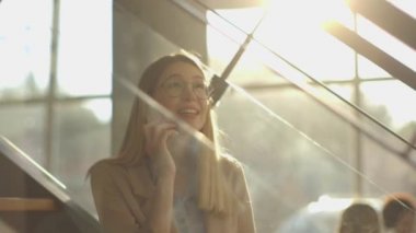 Gülümseyen modern iş kadını ofisin merdivenlerinde oturuyor, defter üzerinde çalışıyor ve cep telefonuyla konuşuyor.