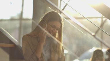 Gülümseyen modern iş kadını ofisin merdivenlerinde oturuyor, defter üzerinde çalışıyor ve cep telefonuyla konuşuyor.