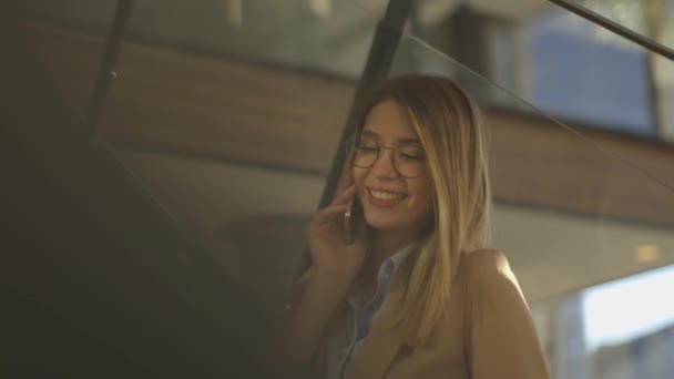 Sorrindo jovem mulher de negócios moderna sentada nas escadas do escritório, trabalhando em notebook e falando no telefone móvel — Vídeo de Stock