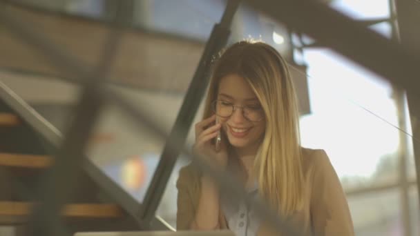 Sorrindo jovem mulher de negócios moderna sentada nas escadas do escritório, trabalhando em notebook e falando no telefone móvel — Vídeo de Stock