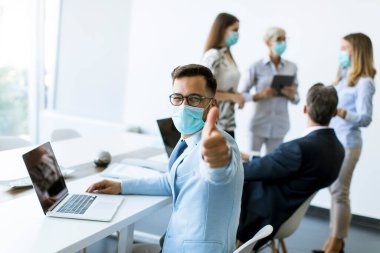 Tıbbi koruyucu maskeli genç iş adamı ofiste dizüstü bilgisayarla çalışıyor ve olumlu başparmak hareketi gösteriyor.