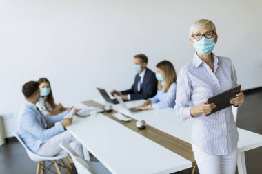 Ofiste dijital tablet kullanan koruyucu maskeli olgun bir iş kadını.