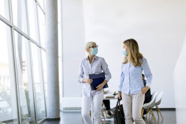 Ofiste yürüyen ve virüs koruması olarak maske takan iki iş kadını.