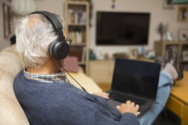 Modern yaşlı bir adam evde kulaklıkla oturuyor ve internetten video görüşmesi yapıyor.