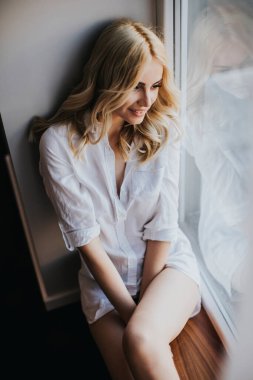 Gün ışığında pencerenin kenarında oturan genç sarışın kadına bakın.