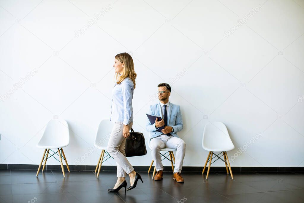 Businesswoman with suitcase passing by young man sitting at chair in the waiting room with a folder in hand before an interview