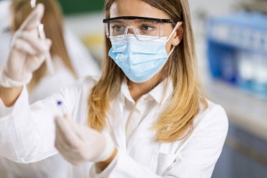 Laboratuvarda koruyucu yüz maskesi ve güvenlik Google 'ı takan kadın doktor şırınga ve ilaç şırıngası şişesini tutuyor.
