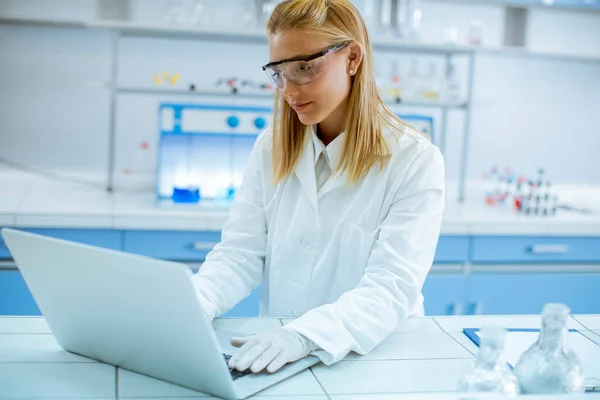 研究室で仕事をしながらノートパソコンを使って白い研究室のコートと保護ゴーグルのかわいい女性研究者 — ストック写真