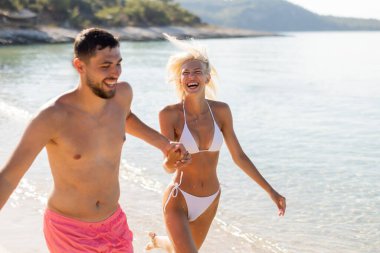 Mutlu bir çift kıyı boyunca el ele koşuyor, gülüyor ve güneşin sıcaklığını kucaklıyor ve güzel bir günde hafif bir meltem esiyor..