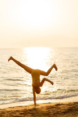 Güneş okyanusun üzerinde batarken bir insan kumlu sahilde neşeli bir takla atar. Sıcak parıltı, yaz eğlencesinin ve özgürlüğün özünü yansıtan huzurlu bir atmosfer yaratıyor..