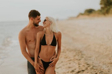 Genç bir çift kumlu bir plajda sarılır, güneş batmaya başladığında kahkahalarını paylaşır, kıyı şeridinde sıcak bir parıltı yayar..