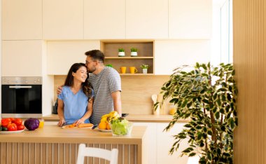 Şık bir mutfakta, bir çift doğal ışıkla çevrili renkli meyve ve sebzelerle sağlıklı bir yemek hazırlarken neşeli bir anı paylaşır..