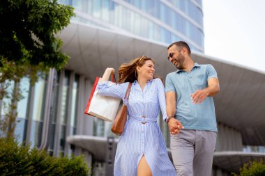 Aydınlık bir günde, bir çift el ele gezer, alışveriş torbaları taşırken gülümser ve güler. Modern mimari, keyifli gezileri için çarpıcı bir zemin görevi görüyor..