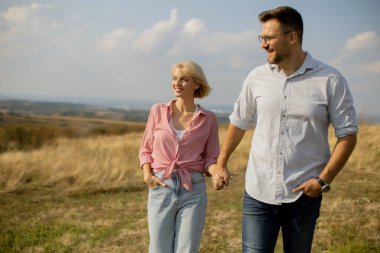 Bir çift, parlak mavi gökyüzünün altındaki yemyeşil bir arazide el ele yürüyor. Mutlu ve rahat görünüyorlar, sıcak havanın ve manzaranın tadını çıkarıyorlar..